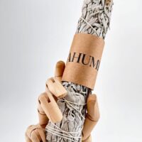 Wooden mannequin hand holds dried sage bundle wrapped and labelled for smudging rituals.