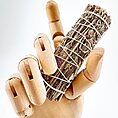 Wooden mannequin hand holding a dried sage bundle for smudging, on a white background.