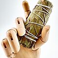 Wooden mannequin hand holding dried eucalyptus and sage leaves bundle with string on white background.