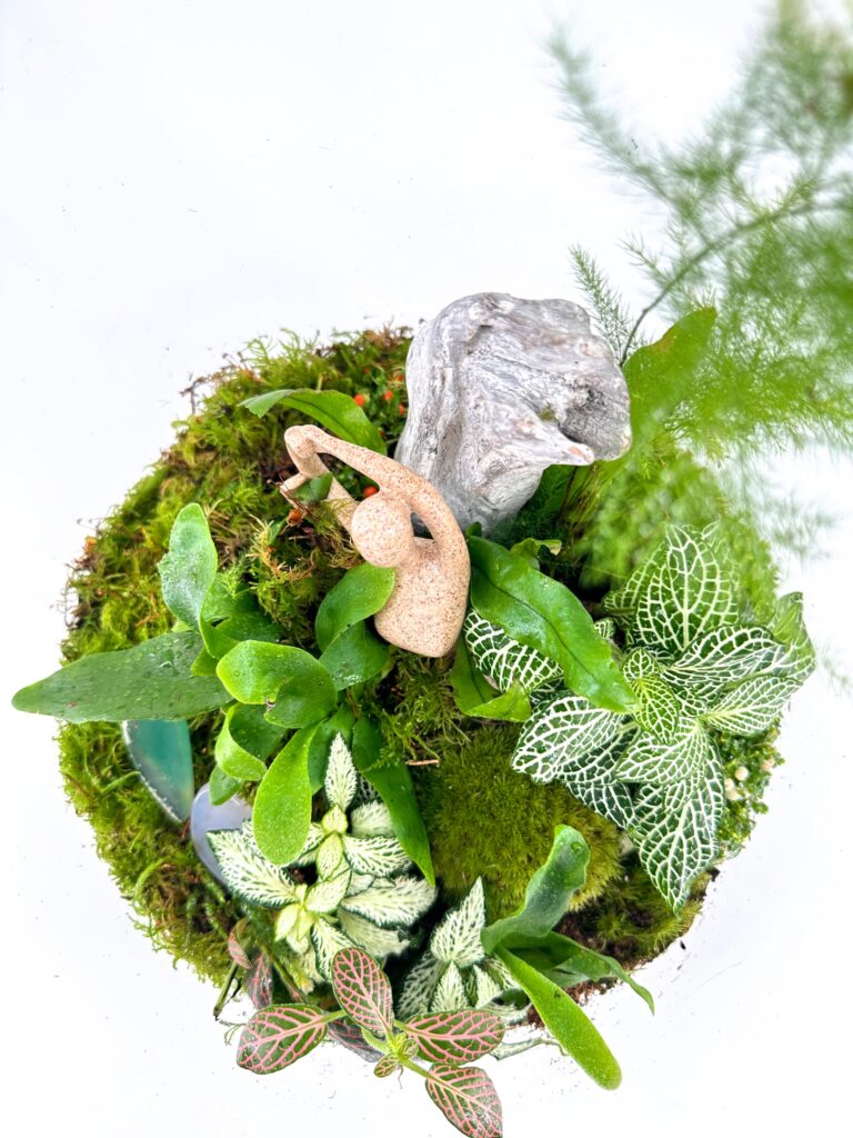 Miniature terrarium with vibrant plants, moss base, driftwood, and an abstract sculpture.