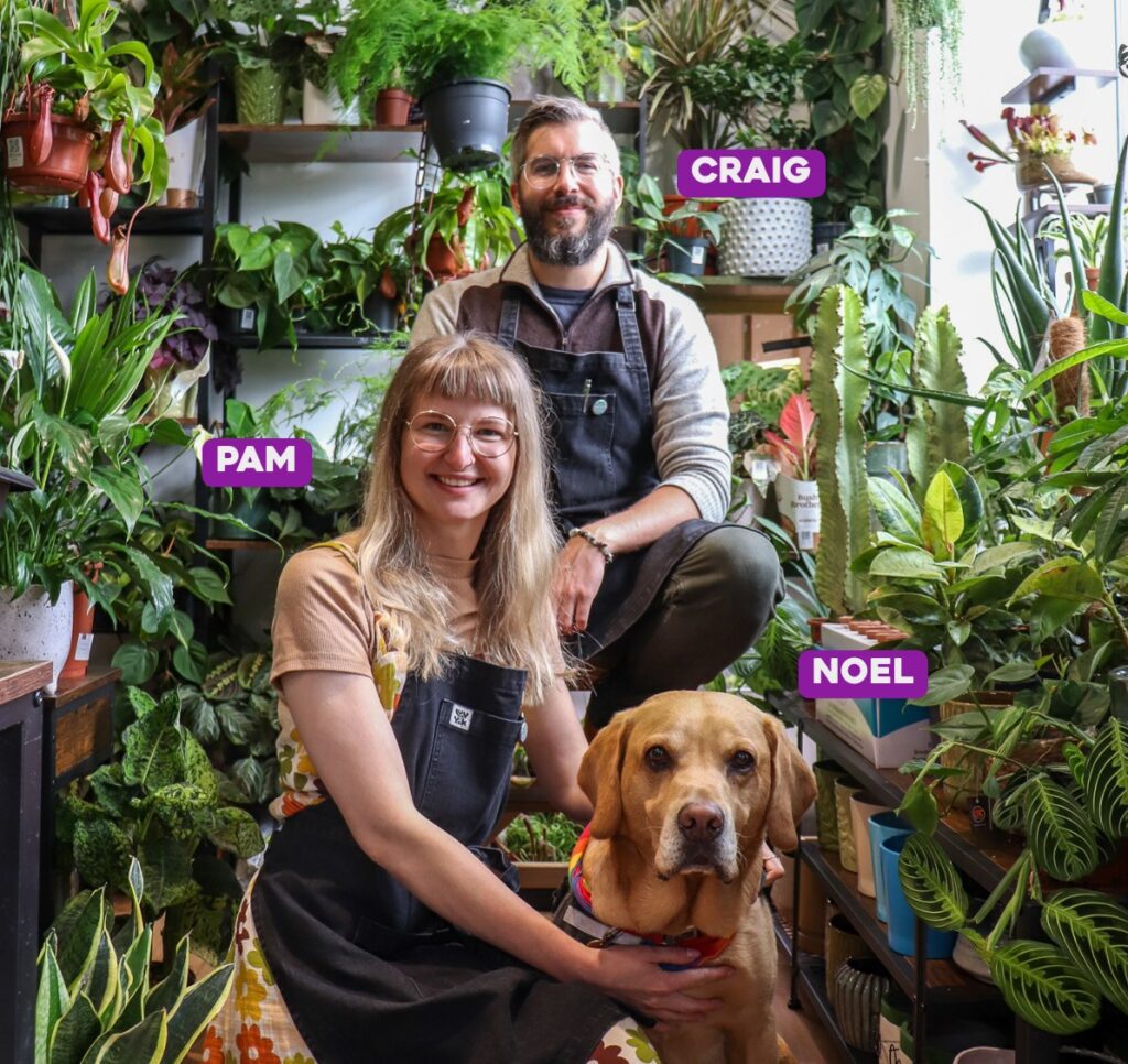 Pam and Craig in lush plant shop with dog Noel, celebrating nature indoors.