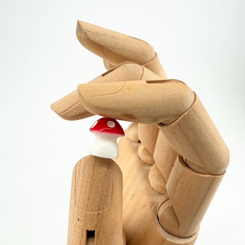 Mini Mushroom Decorations Pack of 3 Surprise Colours A wooden mannequin hand holding a tiny red spotted mushroom decoration between its fingers.